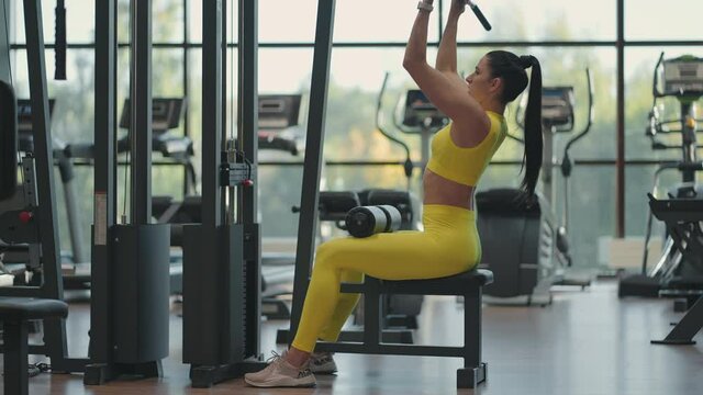 Hispanic woman sitting on a simulator in the gym pulls a metal rope with the weight pumps up the muscles of the back. brunette woman pulls on simulator. performing exercise for back muscles simulator