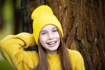 Portrait of modern happy teen girl with dental braces dressed in yellow clothes in park. Pretty teenage girl wearing braces smiling cheerfully. Kid girl in autumn smiling with braces teeth apparatus
