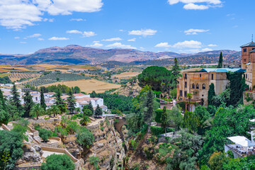 Fototapeta premium Paisaje de edificios antiguos y jardines en los acantilados de una garganta, con fondo de campo con huertas y montañas un día soleado con cielo azul. Desde Ronda, Málaga, Andalucía, España.