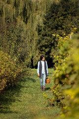 Middle aged woman with dark hair walking on vineyard filed with wicker basket in hands. Female farmer in casual wear and rubber boots spending sunny day for harvesting.