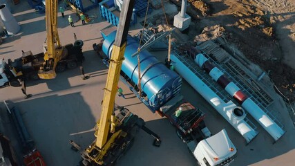 A semi-truck carrying a huge powerful and heavy industrial equipment arrived at its destination - to the construction site of a plant under construction. Large truck cranes unload the delivered cargo
