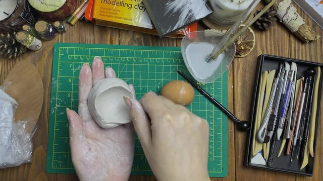 Women's hands makes egg mold out of white clay. Tools for modeling and design of ceramic souvenirs. Hobbies, handmade and crafts. Water, stationery knife, sticks on wooden table.