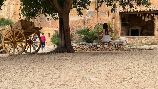 Back view of ittle girl on swing with Sicilian cart  or carretto siciliano at Grotta Mangiapane or Scurati Caves in Sicily, Italy. Slow motion