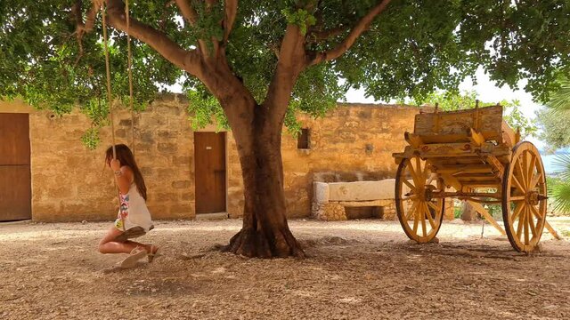 Lttle girl have fun on tree swing with Sicilian cart or carretto siciliano at Grotta Mangiapane or Scurati Caves in Sicily, Italy. Slow-motion