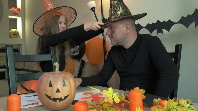 Girl puts a black wizard hat on her dad in decorated room at home. Child and father having fun together, happy family preparing to celebrate Halloween.