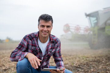 Attractive young farmer holding tablet crouching in front of harvester in soy field