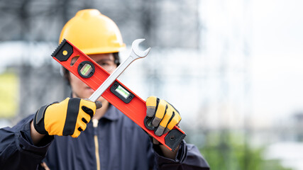 Male Asian mechanic or maintenance worker man wearing protective suit and helmet holding wrench and level tool in cross shape at construction site. Equipment for mechanical engineering project