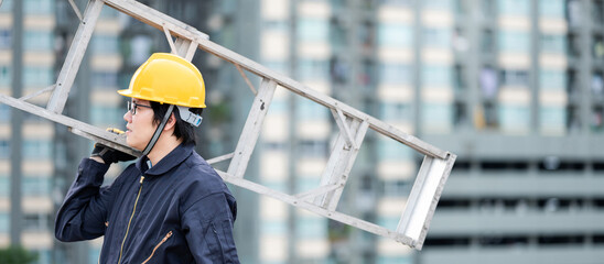 Asian maintenance worker man with protective suit and safety helmet carrying aluminium step ladder at construction site. Civil engineering, Architecture builder and building service concepts