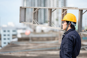 Asian maintenance worker man with protective suit and safety helmet carrying aluminium step ladder at construction site. Civil engineering, Architecture builder and building service concepts