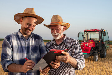 Young farmer and senior agronomist signing documents in field with machinery in background. - obrazy, fototapety, plakaty