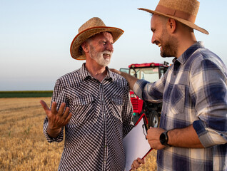 Two male farmers standing in harvested field talking in front of tractor.