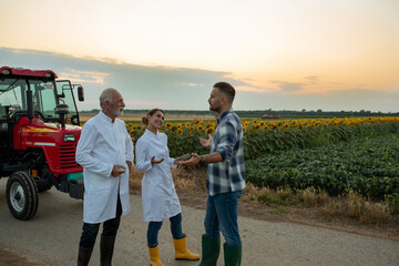 Two scientist speaking with young farmer in front of tractor and sunflower and soy field.