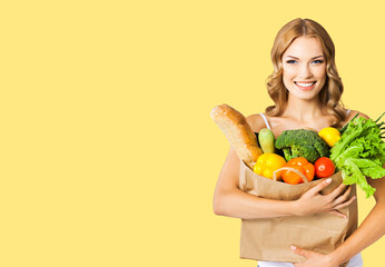 Studio portrait image of happy smiling young beautiful blond woman holding grocery shopping bag with healthy vegetarian food, shopping mall center purchases, isolated over yellow color background