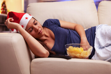 Obraz na płótnie Canvas Exhausted woman falling asleep on couch while watching xmas movie on television during christmastime enjoying winter season in x-mas decorated kitchen. Adult person celebrating christmas holiday