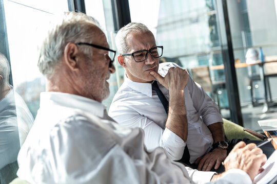 Picture Of Young Business Man Taking To His Older Business Partner. They Are In White Shirt And Black Tie. They Are In A Hotel Lobby. 