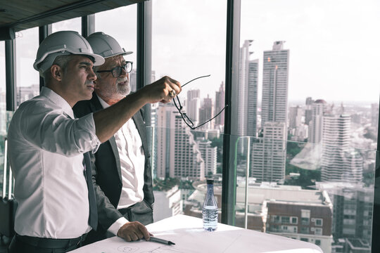Picture of two engineer discussing their construction project together in a hotel lobby. They are wearing white shirt , suit and hardhat. They are drawing on a piece of paper with pen in their hands.