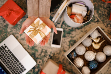 Christmas background with decorations and gift boxes. Woman hands holding wrapped white box with golden bow.