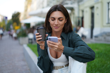 Fototapeta premium Woman with cup of coffee outdoor at city street at sunset happy smiling enjoying summer days look at mobile phone screen