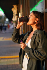 Woman with cup of coffee outdoor at city street at sunset with electronic cigarette catching sun rays enjoying smoking