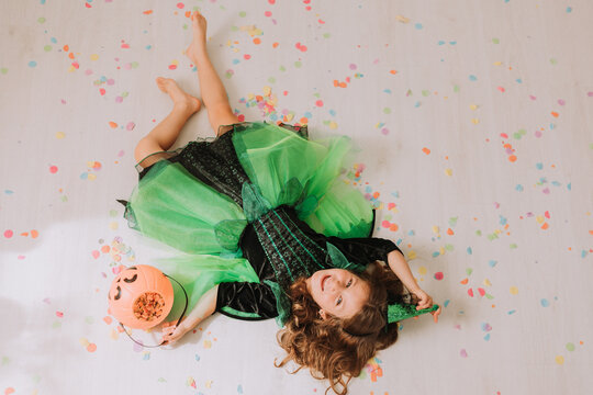 cute little girl in green Halloween costume of a witch or fairy lies on the floor strewn with colorful confetti. top view. pumpkin basket for sweets, lantern jack, happy halloween. autumn concept