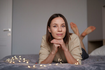 Pretty Caucasian woman smiling with fairy lights laying on bed. Comfy home. Dreaming time