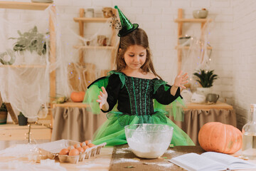cute little girl in a green Halloween costume of a witch or fairy prepares a pumpkin pie, barm brack in a festive decorated kitchen. space for text. High quality photo