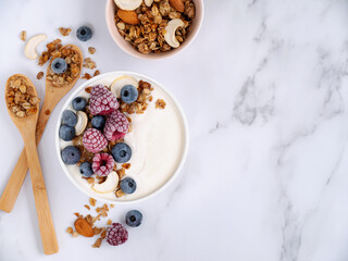 Homemade yogurt with frozen raspberries and blueberries, cashews, granola and almonds with wooden spoons on a light marble table. flat lay. copyspace. Horizontal