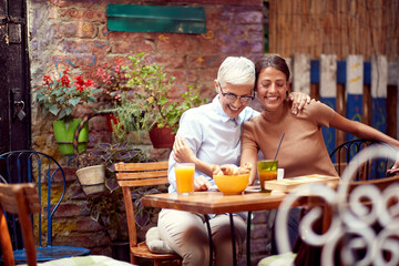 mother and daughter having breakfast together in outdoor cafe, hugging each other, smiling