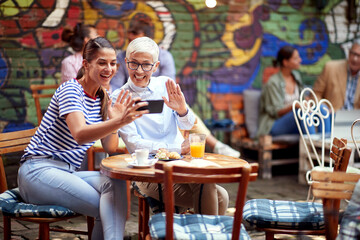 two females, different ages, sitting in outdoor cafe, using cell phone for online communication, talking to somebody, waving.