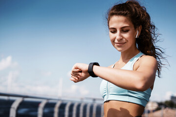 Sports and clothing for women. The calorie counting coach looks at the smartwatch. Fitness break in the city. The athlete leads a healthy lifestyle. Cardio training for weight loss. - obrazy, fototapety, plakaty