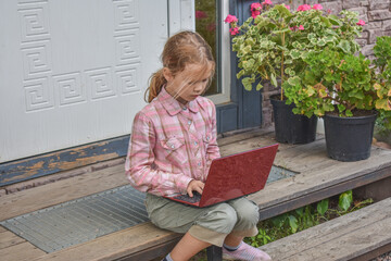 the girl child with a laptop is sitting on the porch of the house during a lesson on a remote photo...