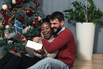 Young happy biracial couple sitting at home in front of the Christmas tree decorated with decorations exchanging gifts for the New Year looking forward to the festive mood - Powered by Adobe