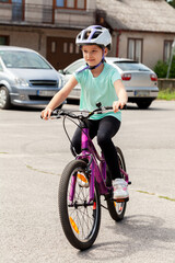 Fototapeta premium Young happy little child, school age girl riding a bike in a bicycle helmet. Outdoor healthy activities, cycling sports and recreation concept, vertical shot, one person. Child learning to ride a bike