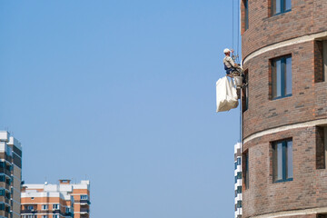Industrial alpinism. A worker in a white helmet hangs on special equipment on the facade of a house under construction. Building construction. copy space. Horizontal photo.