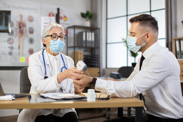 Female medical worker using modern infrared thermometer for checking temperature of caucasian businessman. Doctor and patient wearing protective face mask. Cabinet of modern clinic.