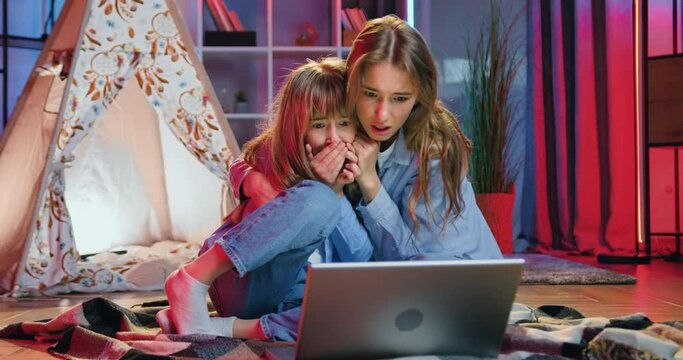 Beautiful scared two sisters,younger and eldest,watching horror on computer and hugging each other from scare and screaming from terrible stages