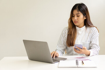 A smiling Asian businesswoman working on a laptop and holding a phone in the office sits at a desk in a financial conference room to check the accuracy of the numbers in the graph document report. fin