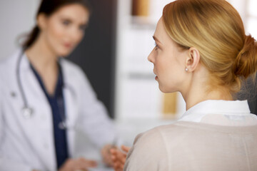 Fototapeta premium Friendly smiling female doctor and patient woman discussing current health examination in sunny clinic. Perfect medical service in hospital. Medicine concept