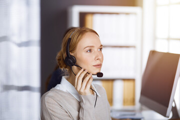 Friendly caucasian businesswoman talking by headset in sunny office. Call center and diverse people group in business