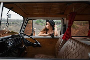 Beautiful girl in a hat with a camera, standing by a retro van, a view from the salon.