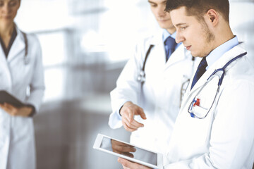 Group of doctors are checking medical names on a computer tablet, with a nurse with a clipboard on the background, standing together in a hospital office. Physicians ready to examine and help patients