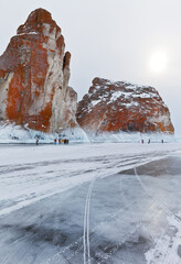 Baikal Lake on a cold February day. A group of tourists travels on the ice of a frozen lake and photographs the red icy cliffs of Olkhon Island. Winter holidays and recreation