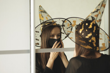 preparing for Halloween in covid,teenage girl at the mirror puts on a protective mask in a witch costume before a holiday party on the eve of all saints
