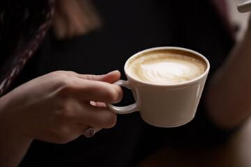 Female hands holding a white cup of delicious coffee with foam over wooden table, side view. Valentines Day coffee