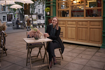 Obraz na płótnie Canvas Beautiful Caucasian female is drinking delicious coffee and looking aside while sitting in the street cafe along and waiting for dessert
