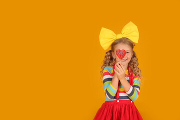 a beautiful blonde girl with a yellow bow in a red skirt, a colored blouse holds a heart-shaped lollipop in her hands