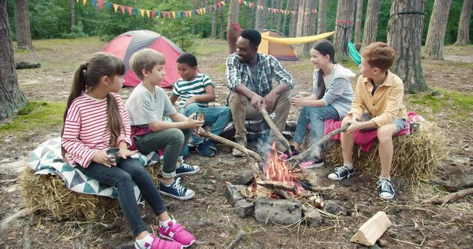 Portrait of caucasian kids and their multiracial teacher having rest time at tent in wood. Little campers relax on sunny day sitting at camping. Spending time in forest concept