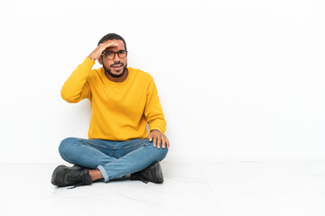 Young Ecuadorian man sitting on the floor isolated on white wall looking far away with hand to look something