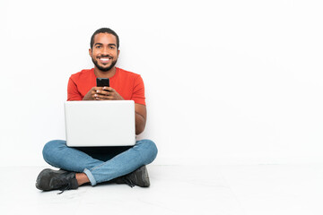 Young Ecuadorian man with a laptop sitting on the floor isolated on white background sending a message with the mobile