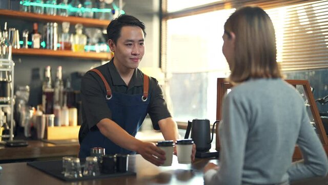 Confidence Asian man waiter or barista working at cafe. Male coffee shop owner preparing takeaway order hot coffee to woman customer on bar counter. Small business entrepreneur food and drink concept.